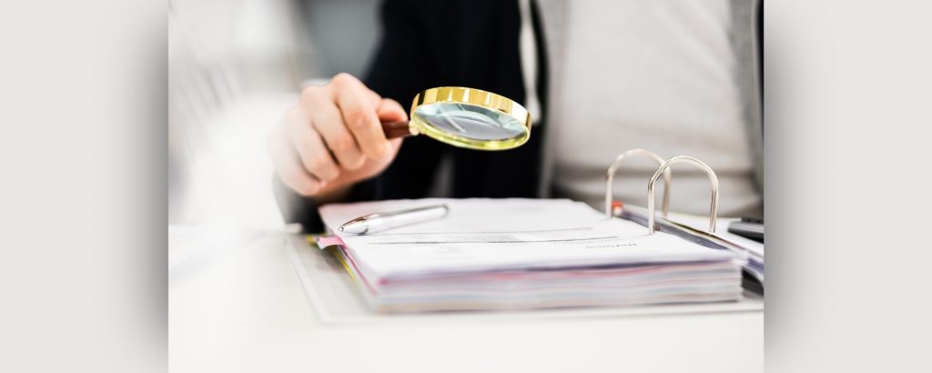 Person holding a magnifying glass over a binder of documents