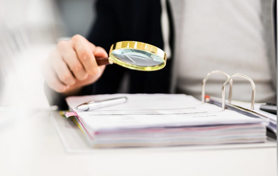 Person holding a magnifying glass over a binder of documents