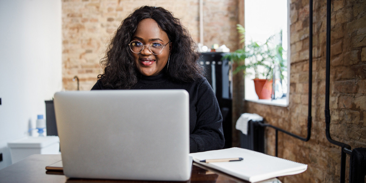 A Black woman with glasses sits at a desk with an open laptop and a notepad.