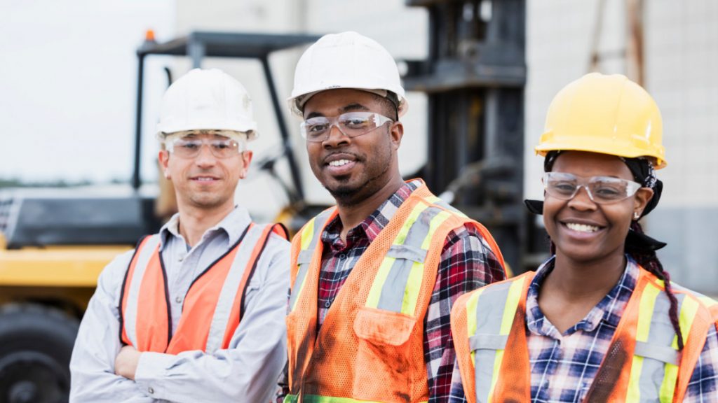 Three people - one white male, one black male, and one black female - all wearing hard hats and orange safety vests stand together smiling.