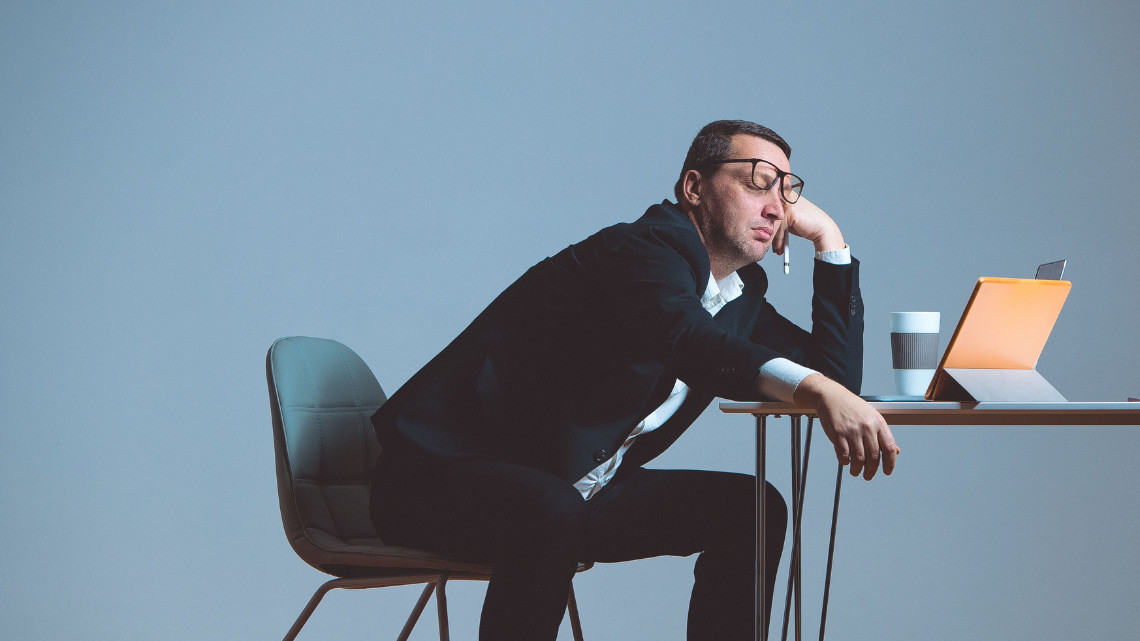 A man in a suit sits slumped over a desk. On the desk sits an open laptop.