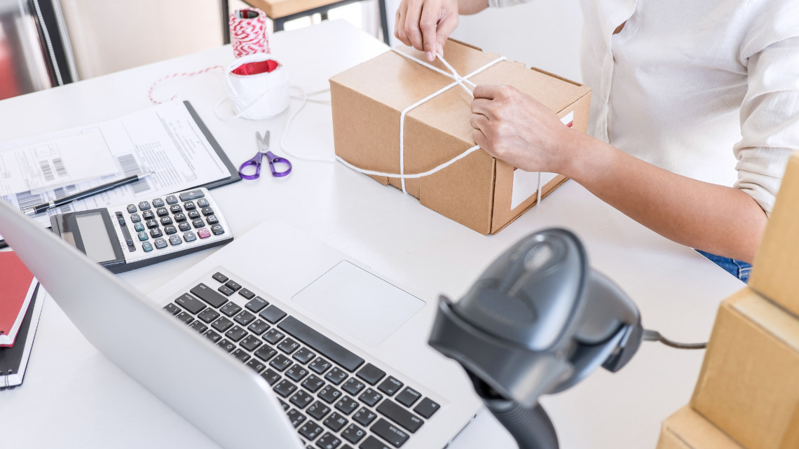 Person tying string around a cardboard box at a shipping workspace with a laptop, calculator, and barcode scanner nearby. Indicating a small business owner or employee.